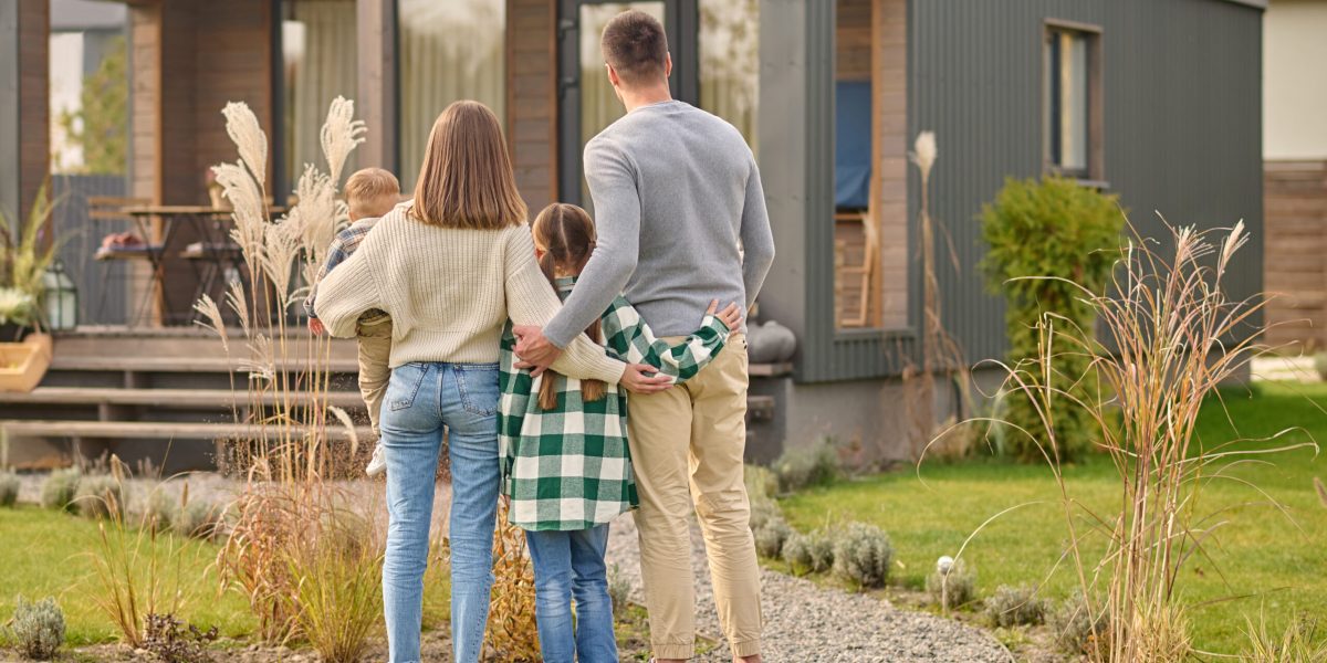 Happy family. View from back of man woman with child and girl standing together hugging and admiring their cozy home on fine autumn day